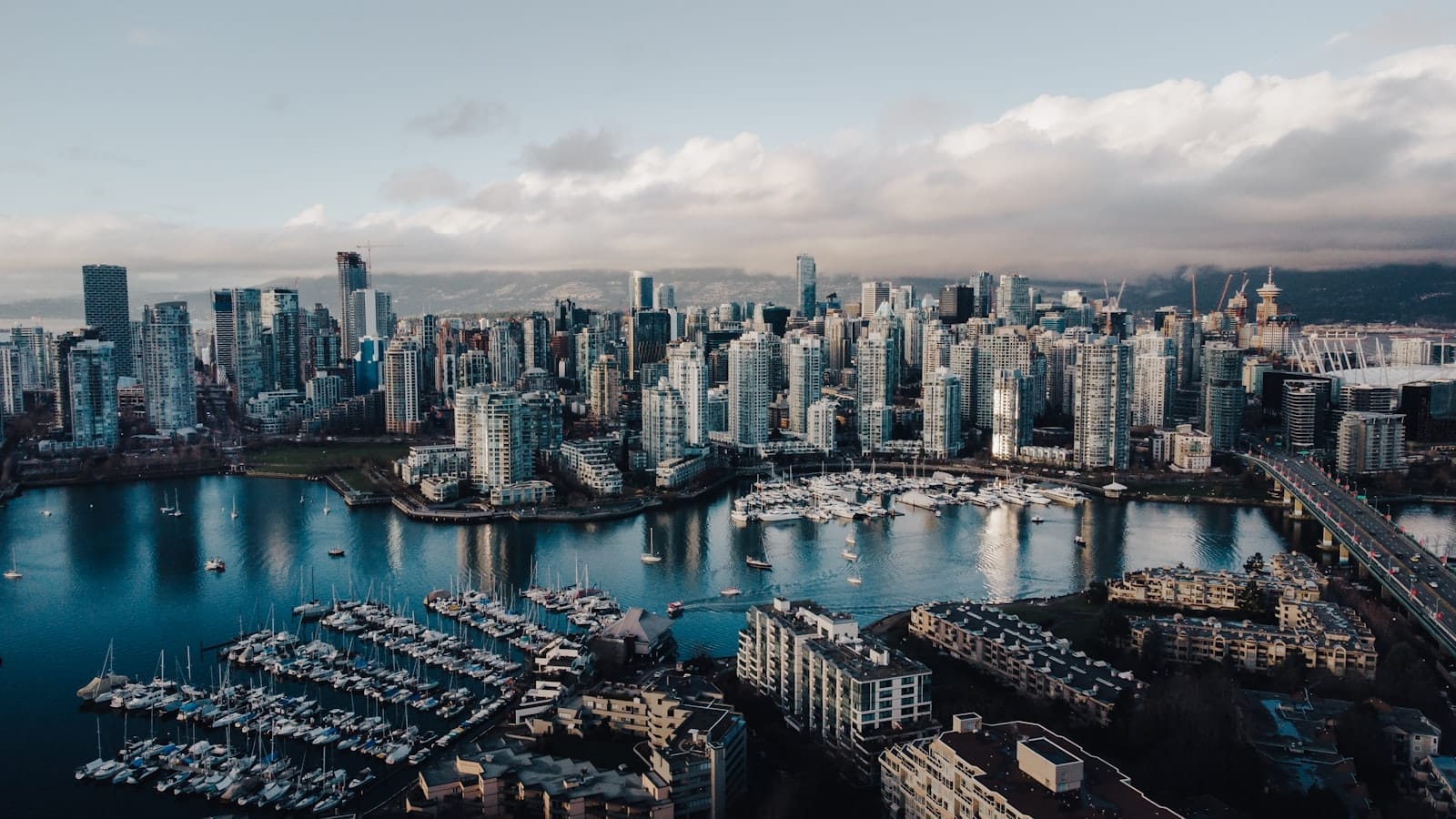 Vista aérea impressionante do litoral de Vancouver, com arranha-céus, iates de luxo na marina e montanhas ao fundo sob um céu nublado.