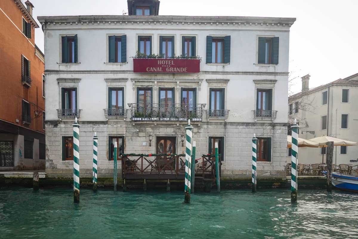 Direkter Blick auf die elegante Kanalfassade des Hotel Canal Grande, mit gestreiften Anlegepfählen und Außenterrasse, die den klassischen venezianischen Luxus widerspiegelt.