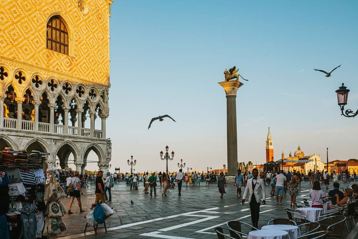 Piazza San Marco in Venedig mit Souvenirständen, Außensitzplätzen, Touristen und ikonischen Säulen bei Sonnenuntergang.