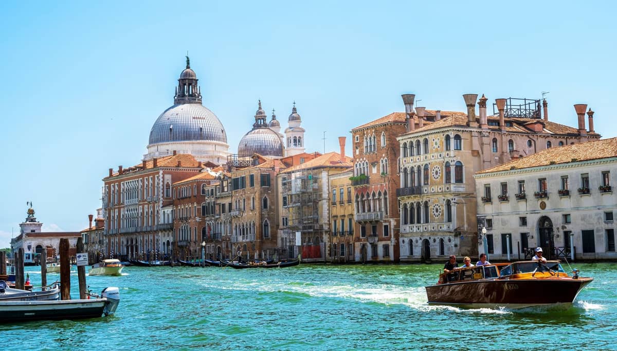 Ein elegantes privates Wassertaxi fährt mit hoher Geschwindigkeit den Canal Grande in Venedig entlang, mit den ikonischen Kuppeln der Santa Maria della Salute und historischen Gebäuden im Hintergrund.