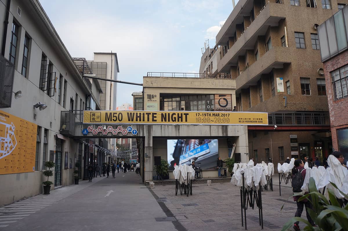 Eingang zum M50 Creative Park in Shanghai mit Besuchern, Industriegebäuden und einem großen gelben Schild mit der Aufschrift M50 White Night