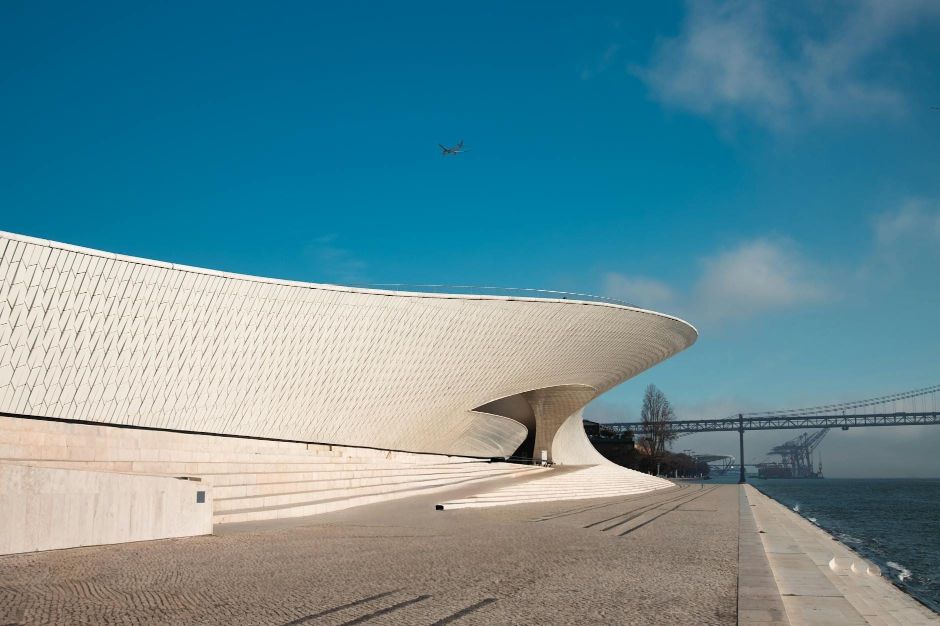 La galleria del MAAT dalla forma ondulata, rivestita di mattonelle bianche, affacciata sulla riva del Tago in piena luce solare, con cielo terso e il ponte sullo sfondo.