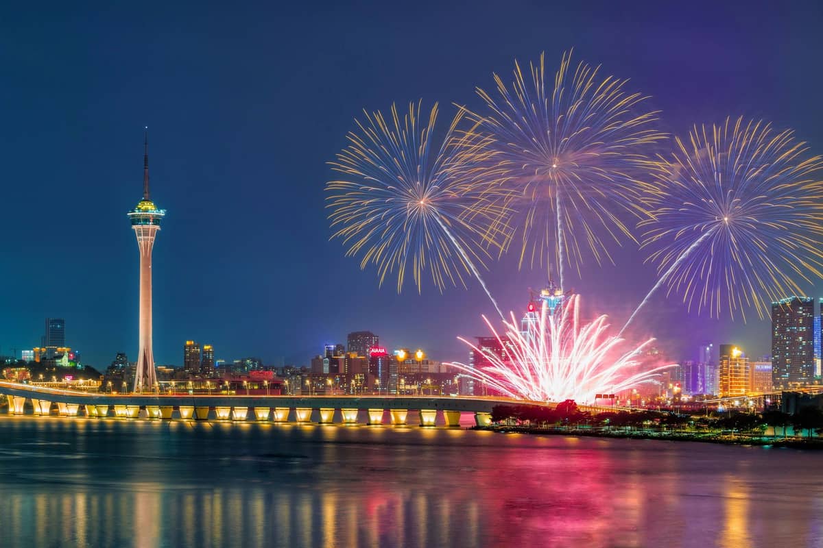Macau Tower und Stadtsilhouette bei Nacht mit Feuerwerk und Spiegelung auf dem Wasser, ein wichtiges Wahrzeichen für Besucher.