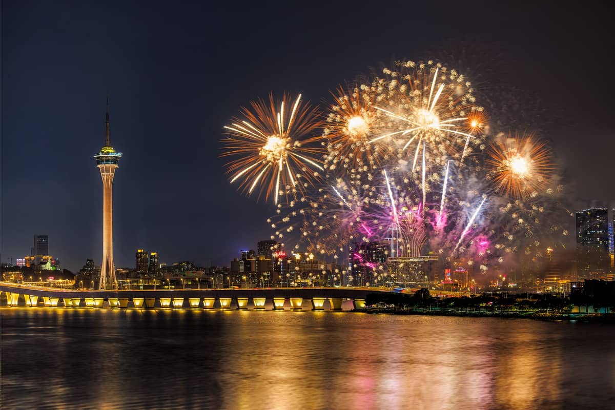 Feuerwerk explodiert bei Nacht über der Skyline von Macau, während sich der Macau Tower und eine beleuchtete Brücke während der Chinesisch-Neujahrs-Feiern im Perlfluss spiegeln.
