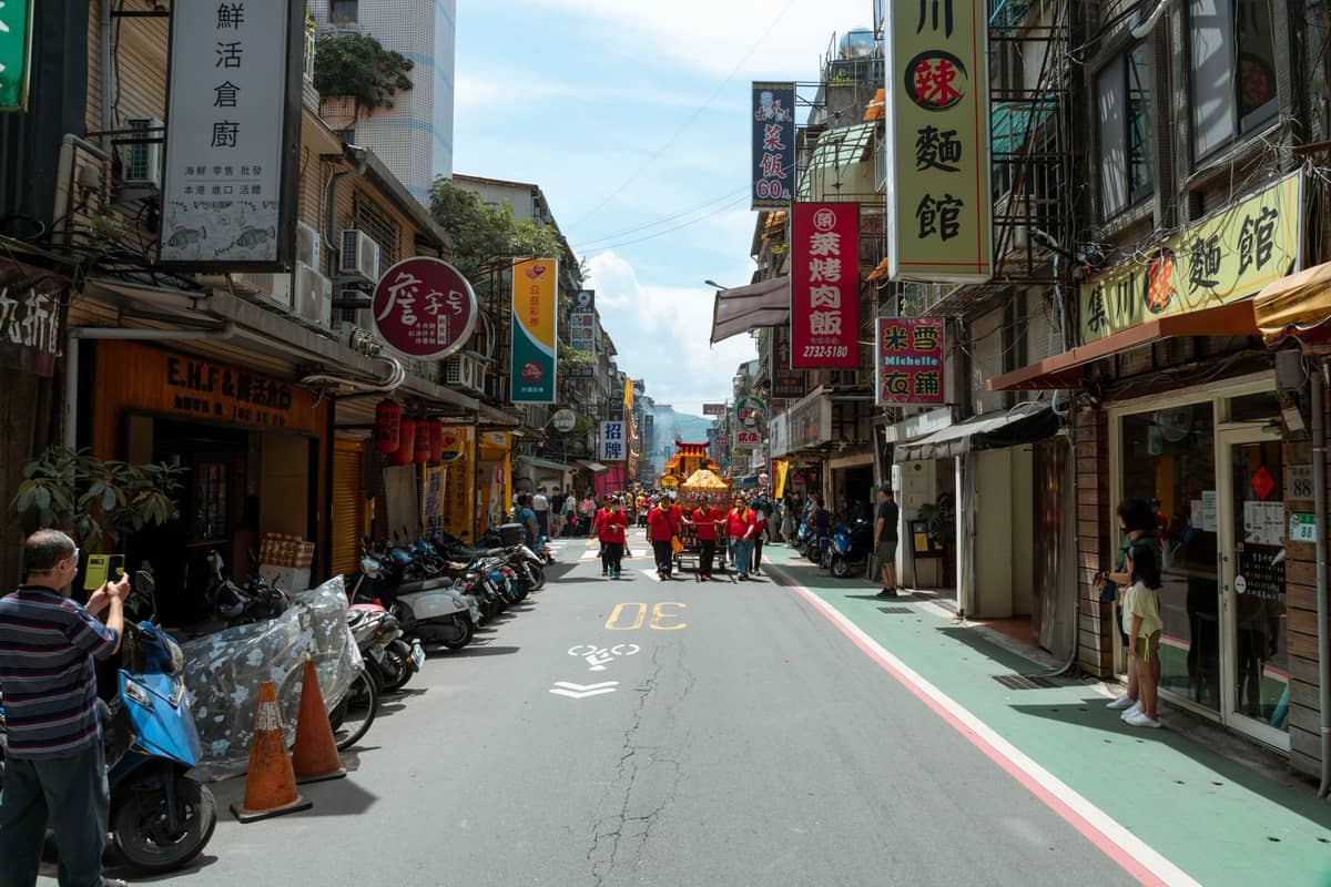 Menschen gehen eine enge Straße in einer asiatischen Stadt mit chinesischen Schildern entlang, einige sind für eine Parade oder ein Festival gekleidet; Motorräder sind am Gehweg geparkt.