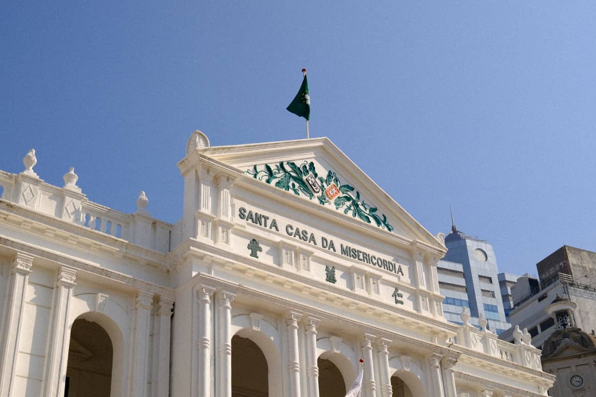 Weißes Bürgergebäude aus der Kolonialzeit mit Bogenfenstern und grüner Flagge, beschriftet mit 'Santa Casa da Misericórdia', unter einem klaren blauen Himmel in Macau.