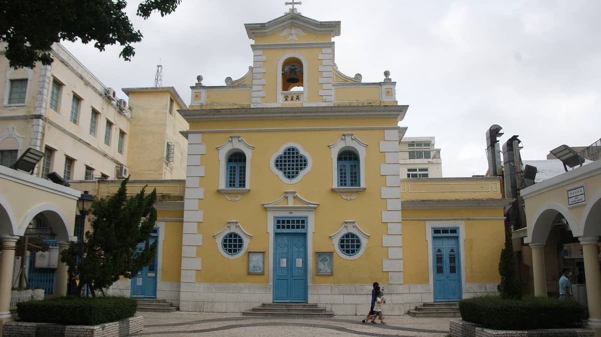 Vorderansicht der gelben Kapelle des Heiligen Franz Xaver mit blauen Türen und Fenstern in Coloane Village, Macau, an einem bewölkten Tag.