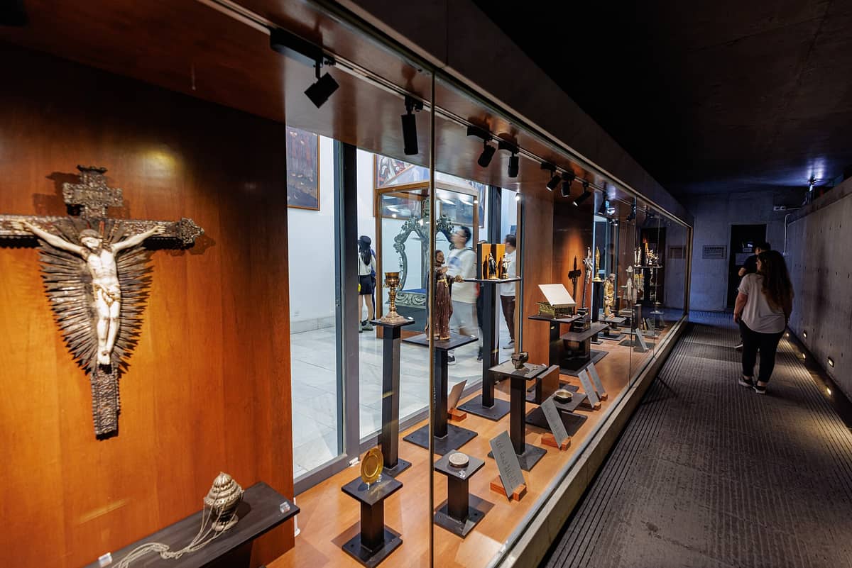 Gläserne Museumsvitrine in Macau mit katholischem Kruzifix, Kelchen und religiösen Reliquien, während Besucher sakrale Kunst und Artefakte betrachten.