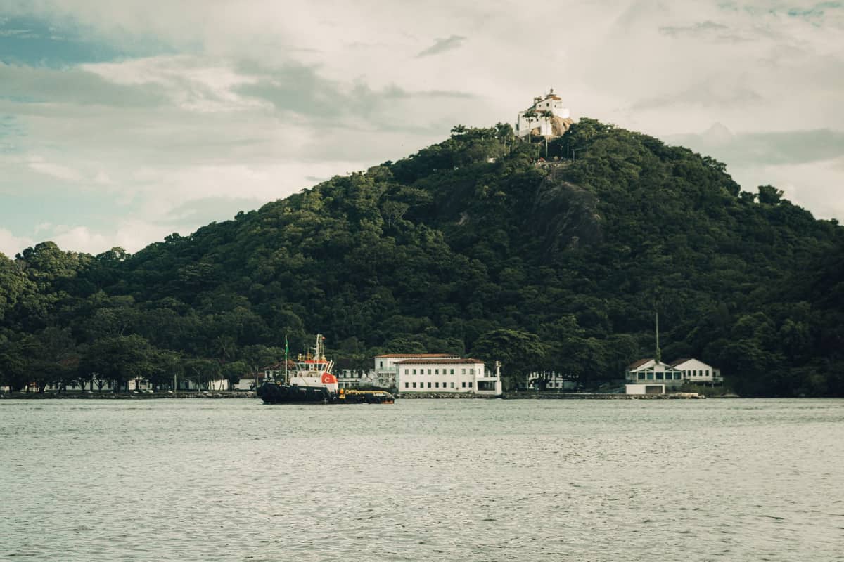 Kleine weiße Kapelle auf einem grünen Hügel, vom anderen Ufer aus gesehen mit Gebäuden und einem Boot im Vordergrund unter einem bewölkten Himmel