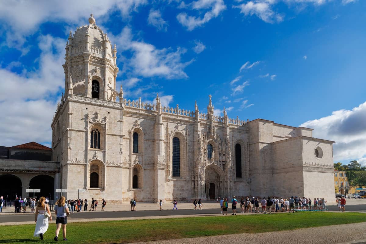 Weitwinkelsicht auf die kunstvolle Steinfassade des Jerónimos-Klosters unter blauem Himmel, mit Touristen, die vor dem historischen Wahrzeichen Lissabons spazieren und sich versammeln.