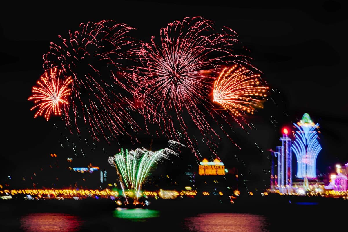 花火สว่างจ้าระเบิดเหนือเส้นขอบฟ้าของมาเก๊าในยามค่ำคืน พร้อมด้วย Grand Lisboa และแสงไฟของเมืองที่สะท้อนบนผิวน้ำ