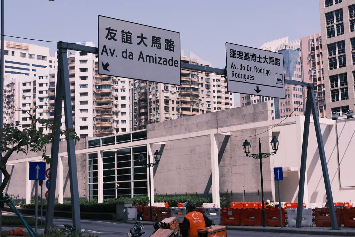 ฉากถนนในมาเก๊าที่แสดงป้ายจราจรของ Av. da Amizade และ Av. do Dr. Rodrigo Rodrigues พร้อมตึกสูงในพื้นหลัง