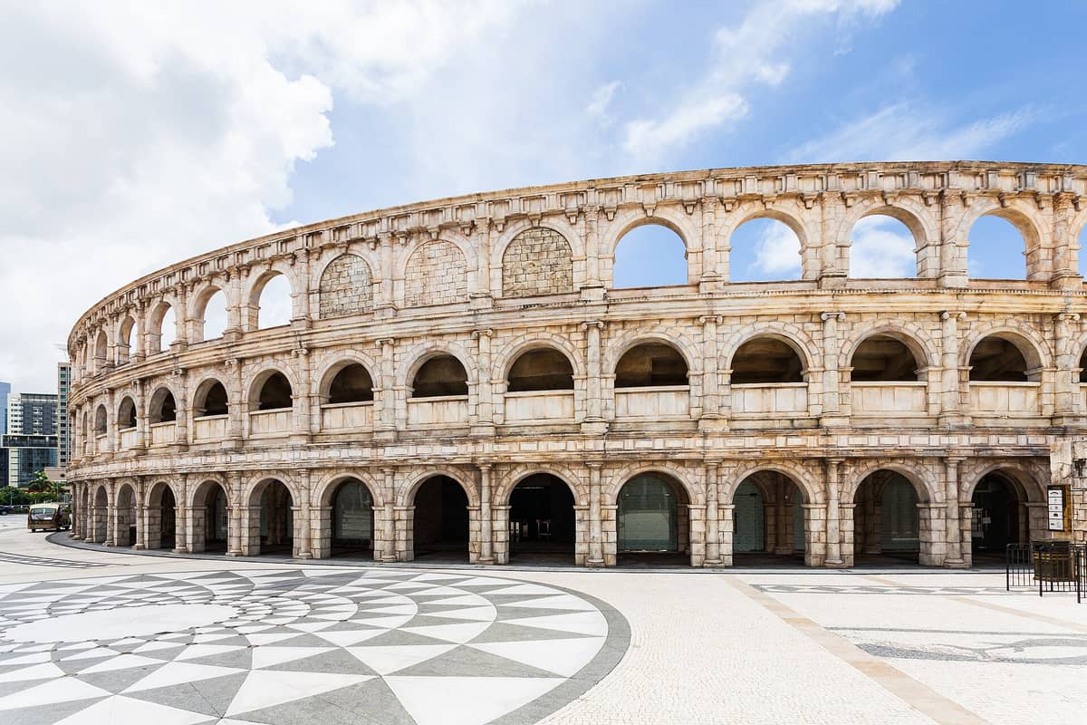 Weitwinkelaufnahme des Amphitheater-Replikats im Macau Fisherman's Wharf: eine Bogenfassade aus Stein und ein gemusterter Platz unter blauem Himmel.