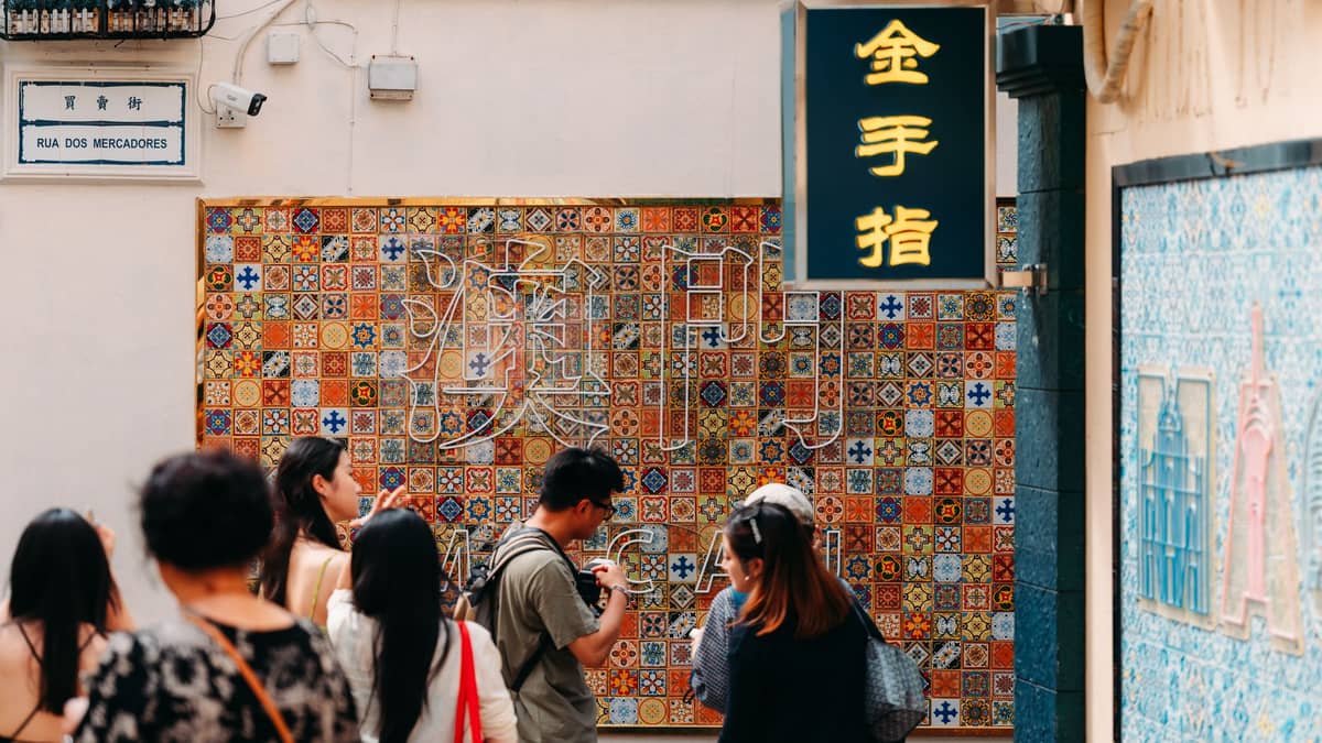 Fußgänger im historischen Macau passieren eine bunte Kachelwand und Ladenchilder in einer engen Straße in der Nähe der Rua dos Mercadores.