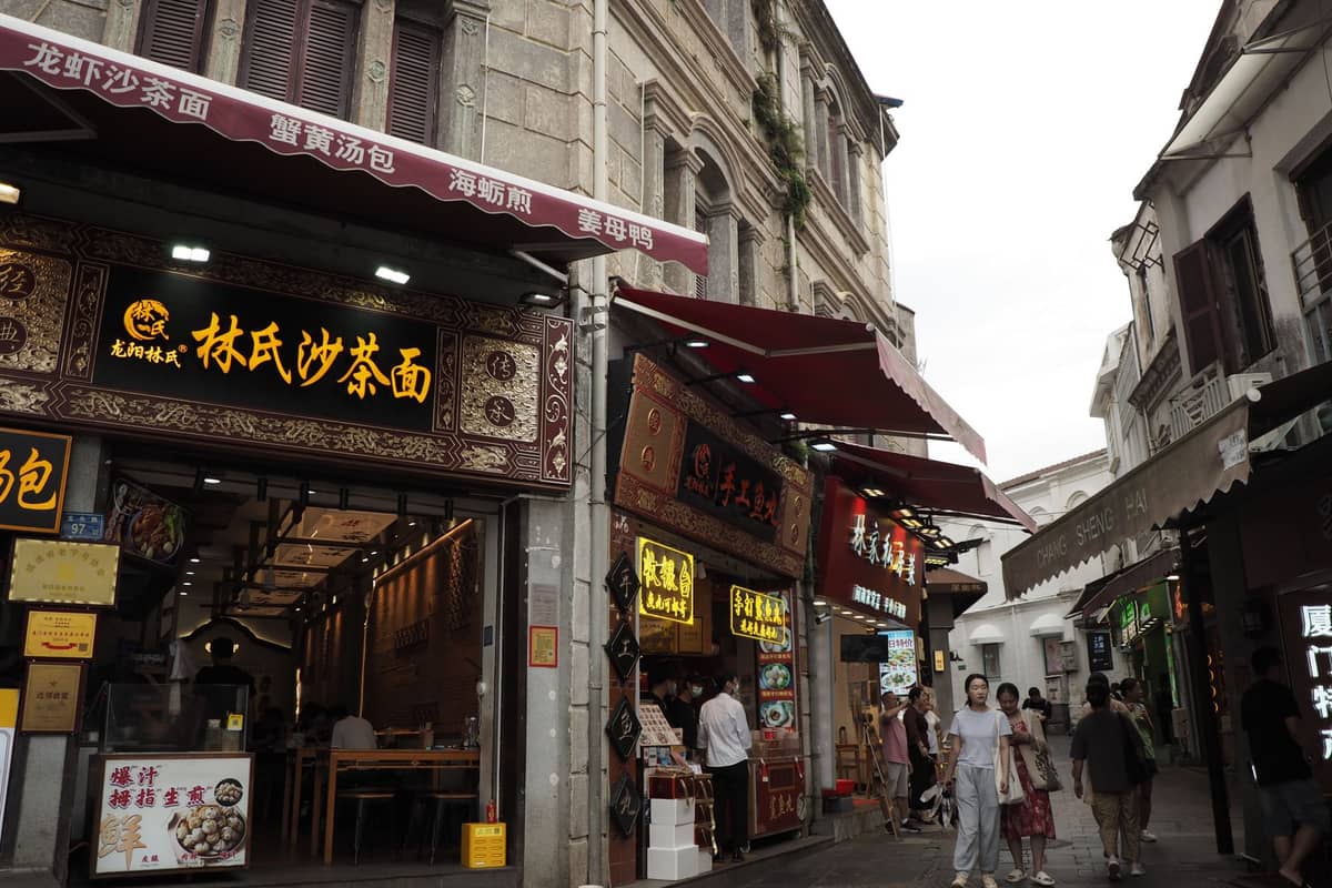 Straßenansicht belebter lokaler Restaurants in einer Fußgängergasse in Macau mit vorbeigehenden Menschen und großen traditionellen chinesischen Restaurantschildern.