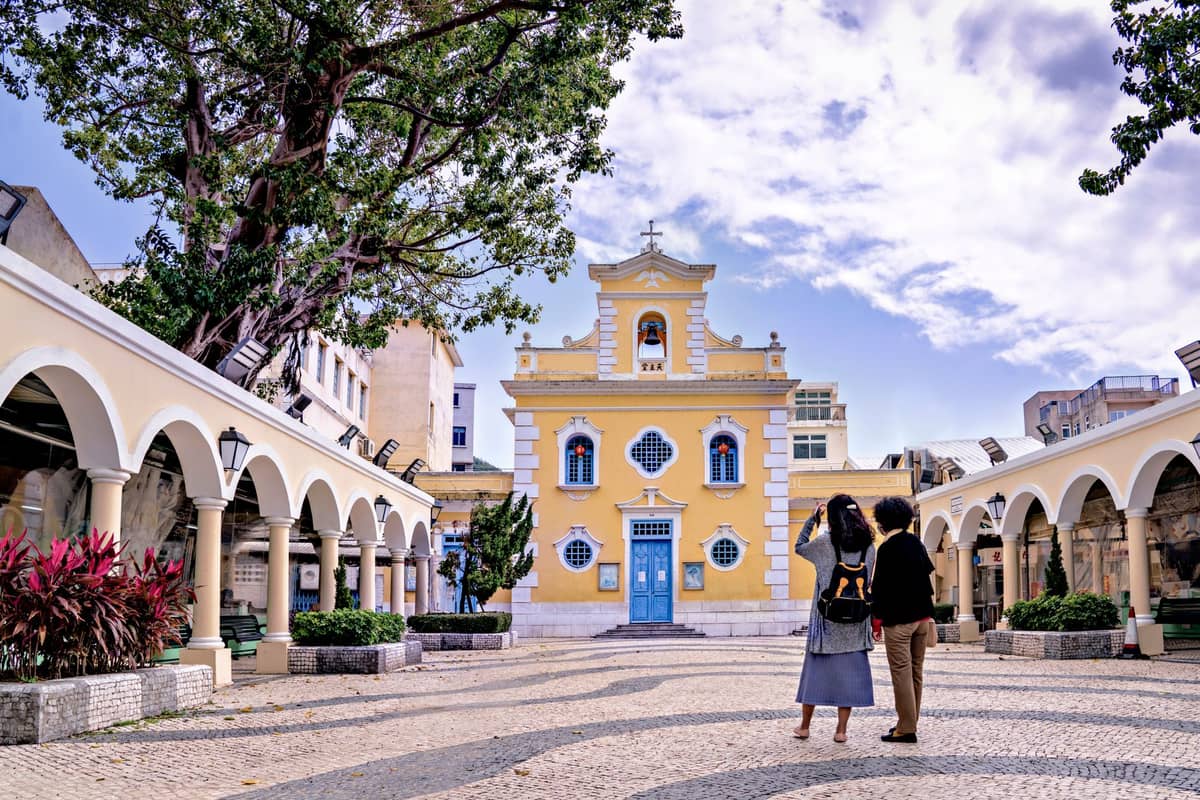Ein Pärchen läuft an einem hellen, angenehmen Tag auf die pastellgelbe St. Dominic's Church in Macau zu.