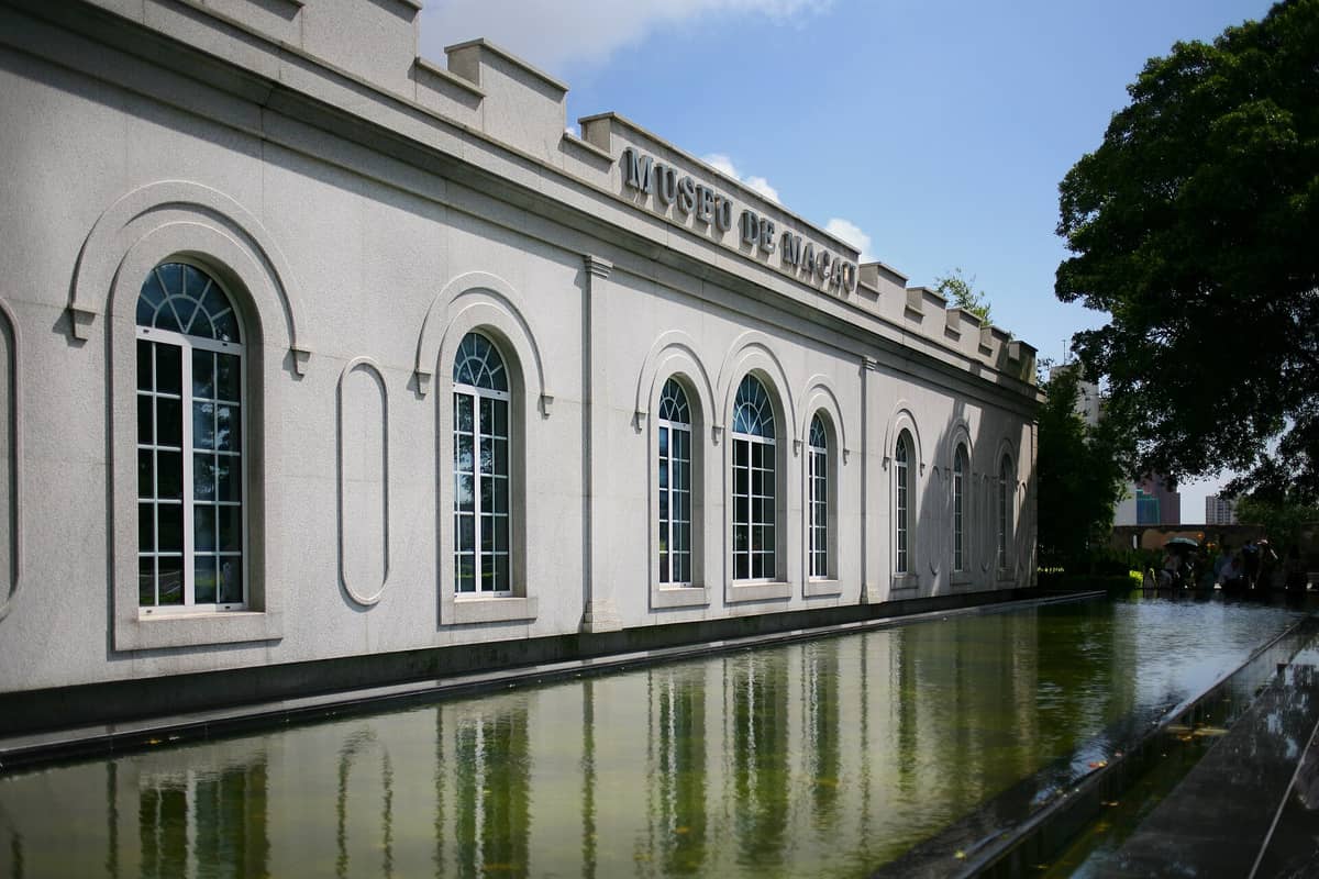 Außenansicht des Macao Museums mit Bogenfenstern und Spiegelung in einem Teich, unter klarem blauem Himmel und umgeben von Grün.
