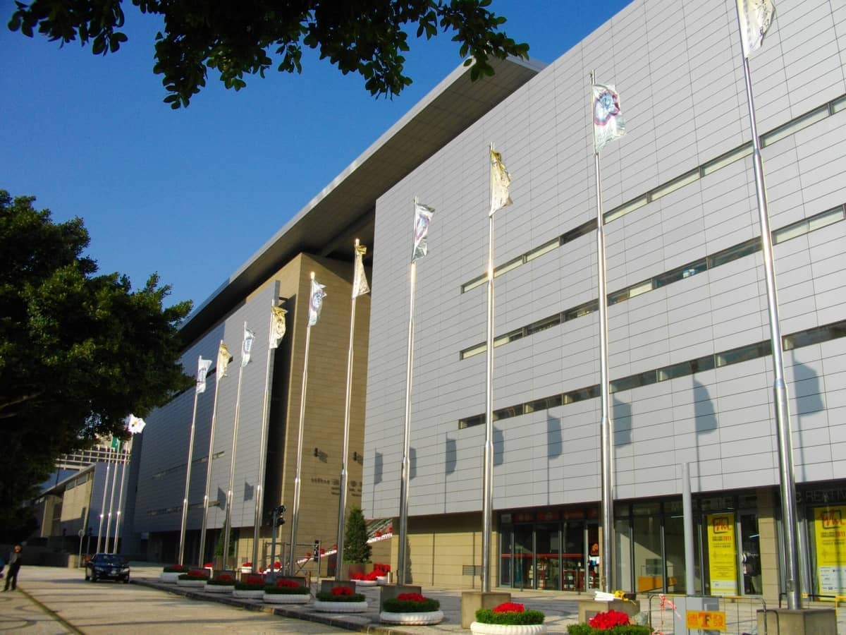 Moderne Außenansicht des Macao Museum of Art mit schlanker Fassade, Fahnenmasten, Blumenkübeln und klarem blauem Himmel.