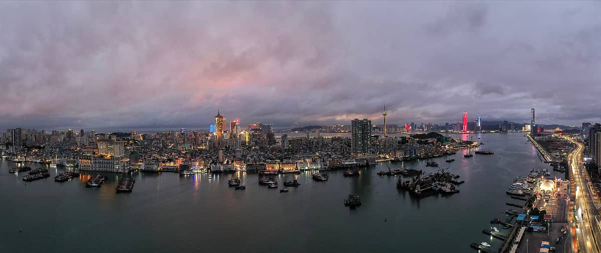 Panoramablick aus der Luft auf die Halbinsel Macau in der Abenddämmerung mit dichter Stadtsilhouette, Uferpromenade, historischen und modernen Gebäuden, dem Macau Tower und Booten im Hafen.