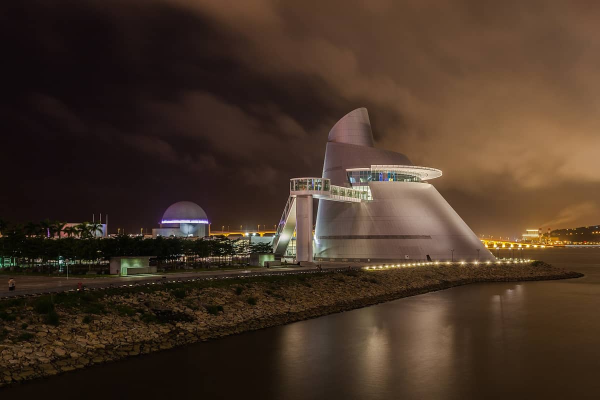 Das markante kegelförmige Silbergebäude des Macao Science Centers bei Nacht, mit der Planetariumskuppel und der beleuchteten Uferpromenade unter einem dramatischen Himmel.