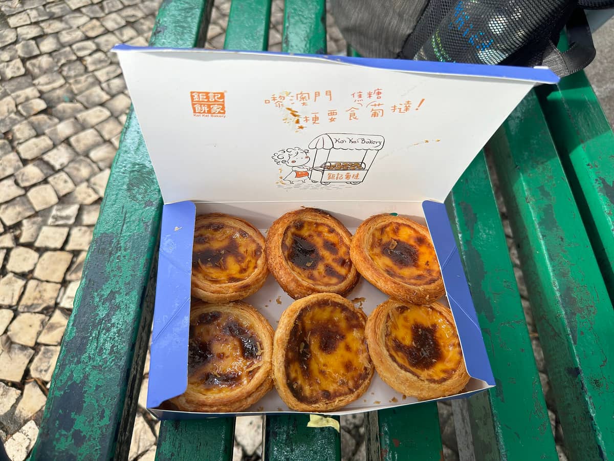 Schachtel mit sechs Macau-Eggentarts von Ko Kei Bakery auf einer grünen Bank mit macaoesischem Kopfsteinpflaster im Hintergrund.