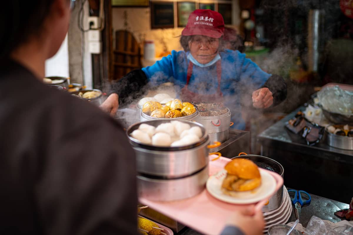 Ein belebter Straßenimbissstand in Macao mit einem Verkäufer, der gedämpfte Brötchen und andere Snacks serviert, mit Körben und Tellern in einer lebhaften Umgebung.