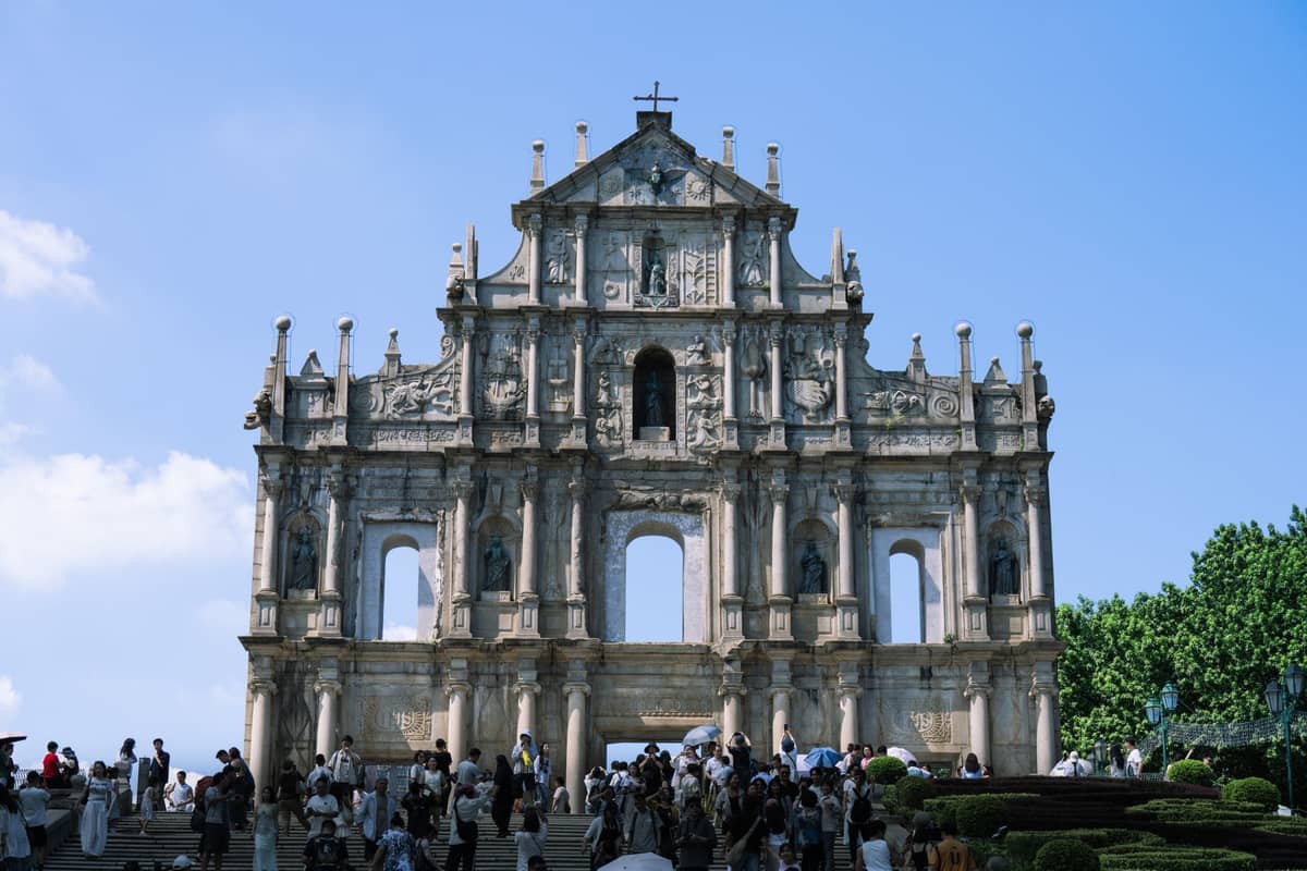 Menschenmassen versammeln sich auf den breiten Steinstufen vor den ikonischen Ruinen von St. Paul's, einem UNESCO-Weltkulturerbe in Macau, unter einem klaren blauen Himmel.
