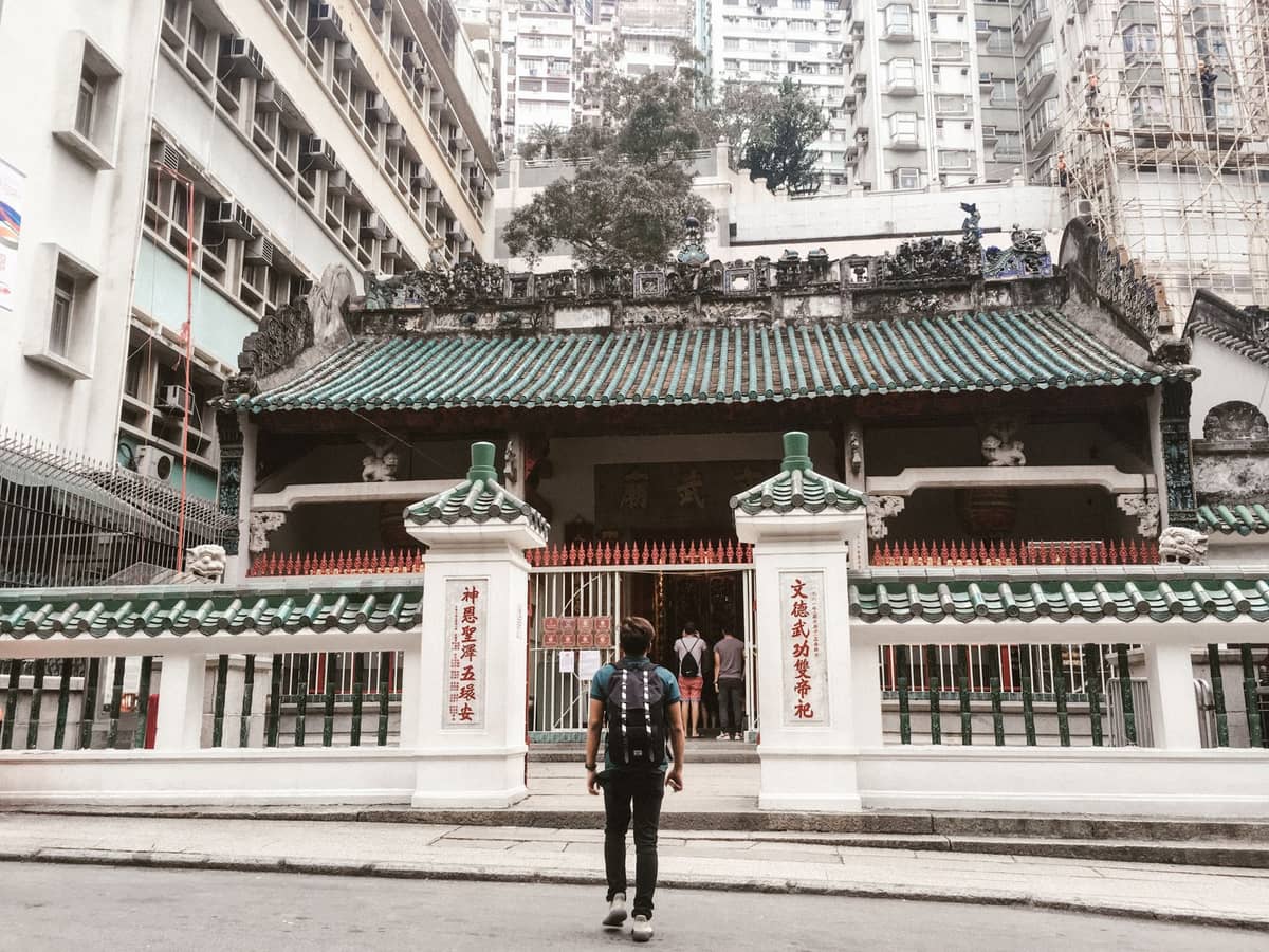 Person vor dem grün gekachelten Eingang eines traditionellen chinesischen Tempels in Macau, umgeben von modernen Wohnhäusern.