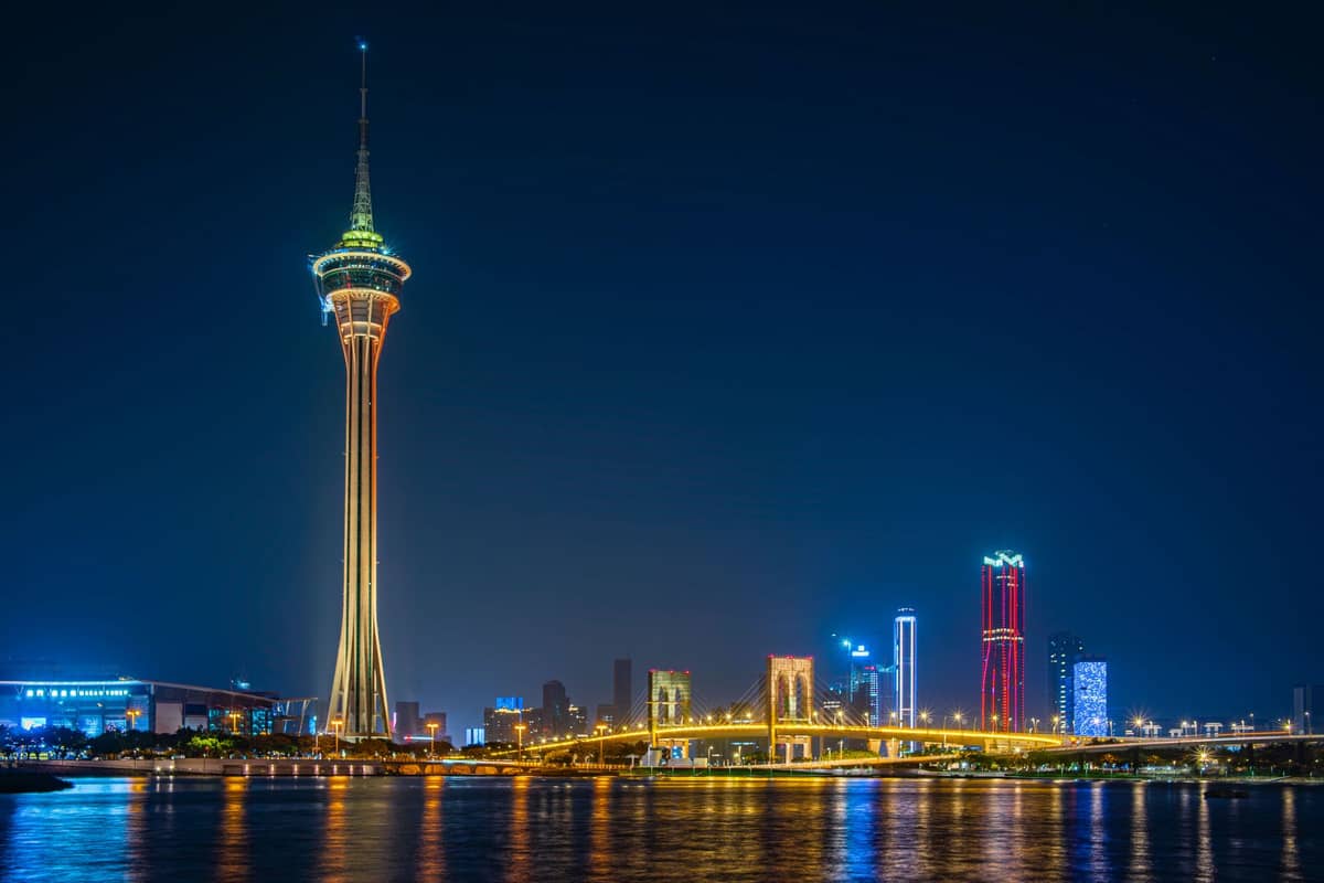 Der Macau Tower leuchtet nachts hell erleuchtet mit Spiegelungen im Sai Van Lake und den Wolkenkratzern Macaus im Hintergrund.