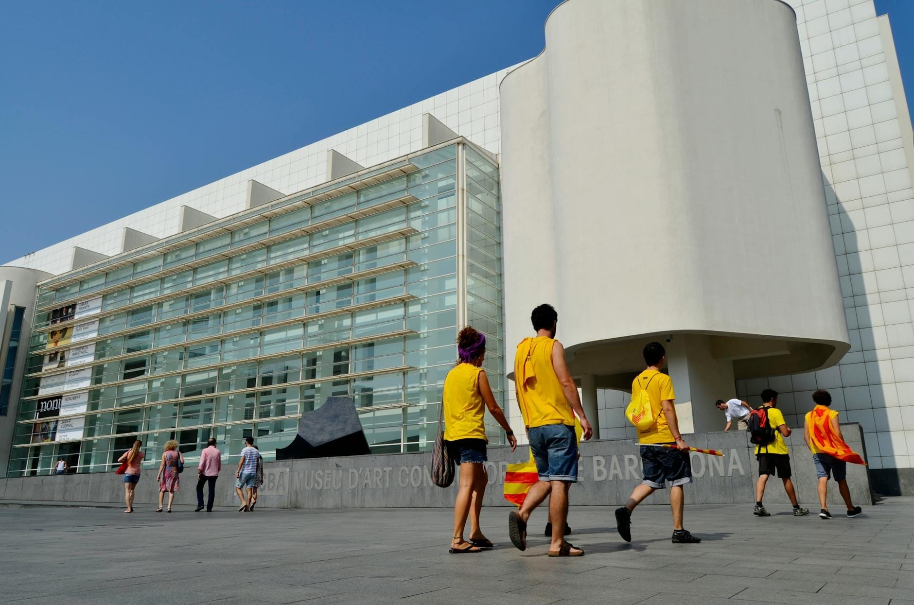 The exterior of MACBA in Barcelona, showcasing the modern white facade, glass walls, and groups of people walking in the sunny plaza.