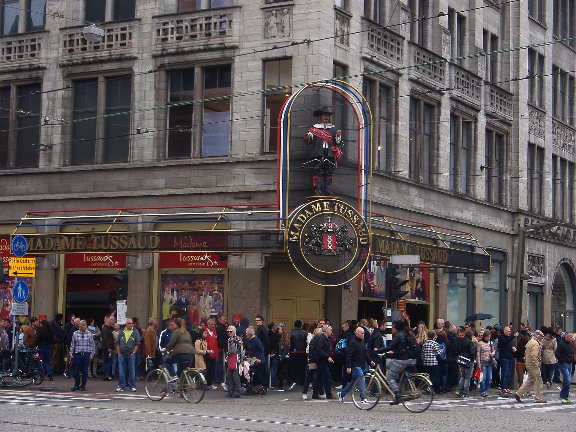 Multitudes esperan fuera de la entrada de Madame Tussauds Amsterdam, un edificio histórico con grandes ventanas y un prominente cartel de figuras de cera sobre la puerta.