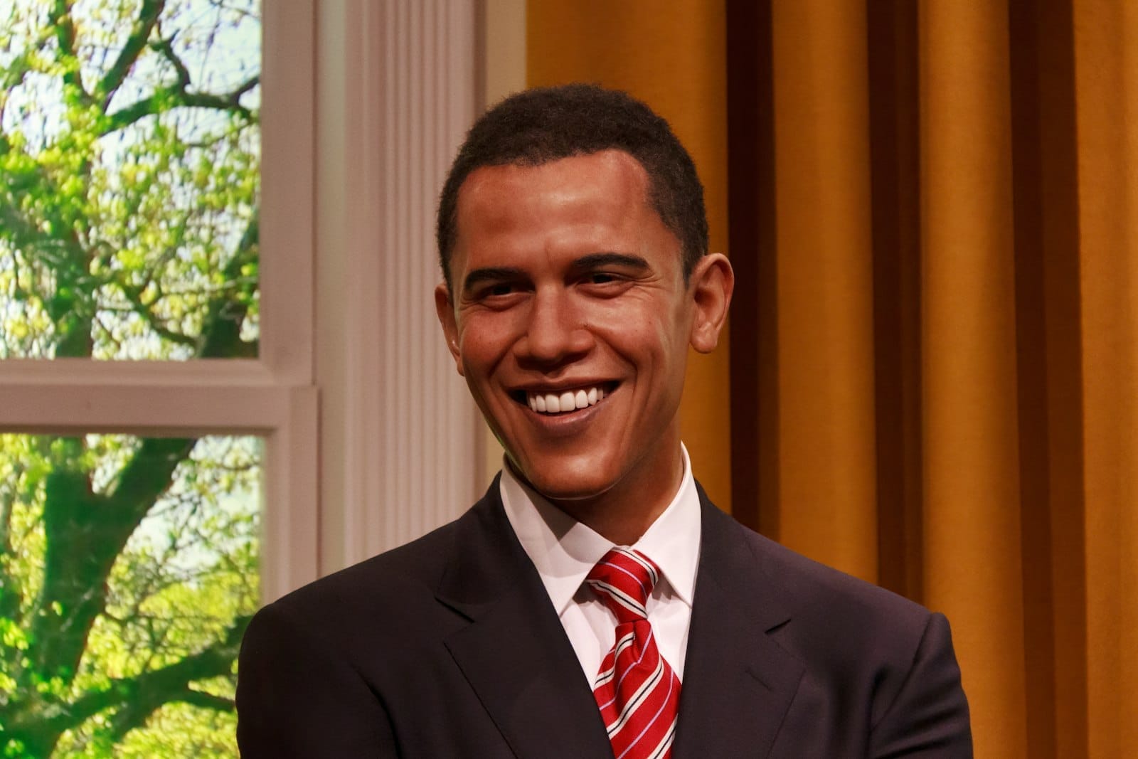 Figura di cera di Barack Obama sorridente da Madame Tussauds London, con tende dorate e una finestra dietro di lui.
