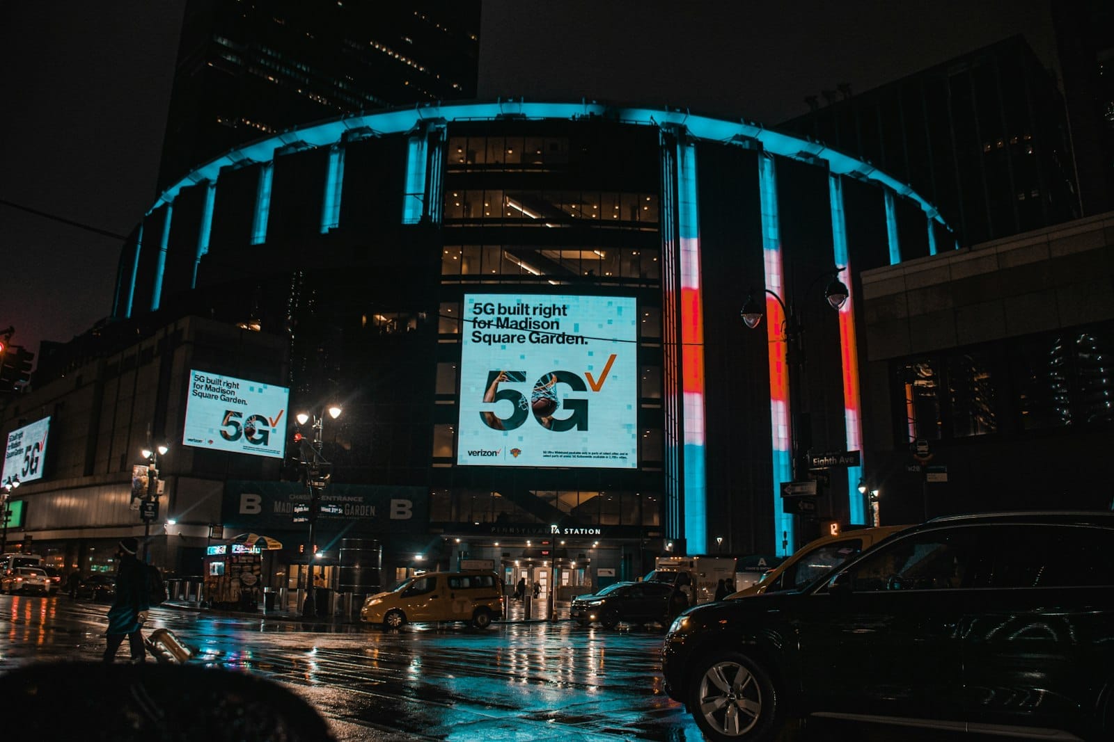 Esterno del Madison Square Garden di notte con luci al neon colorate, cartelloni digitali, strade bagnate e auto a Midtown Manhattan.