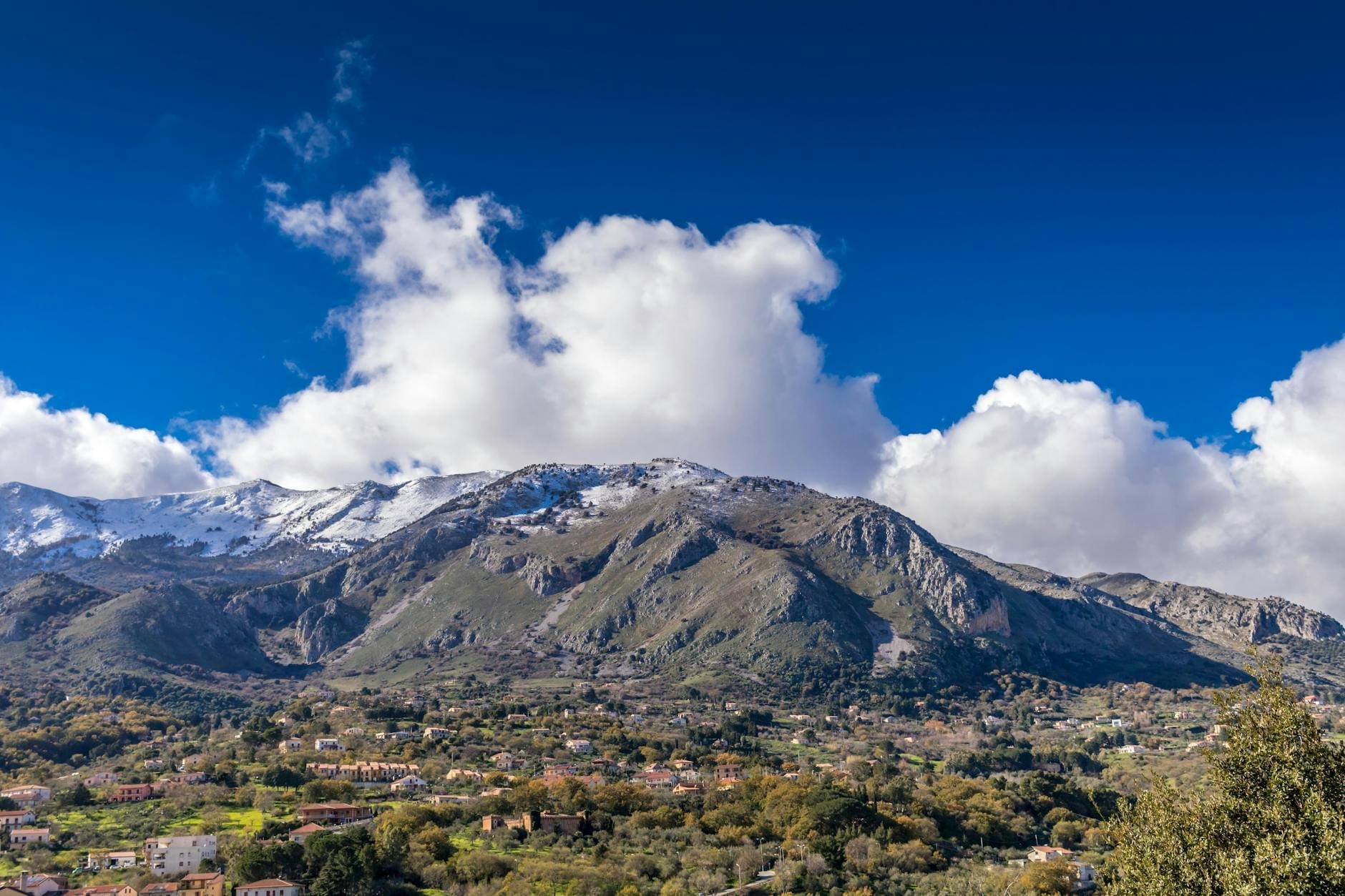 Panoramablick auf schroffe Berge mit schneebedeckten Gipfeln, grüne Täler, verstreute mittelalterliche Ortschaften und dramatische Wolken unter einem leuchtend blauen Himmel im Inneren Siziliens.