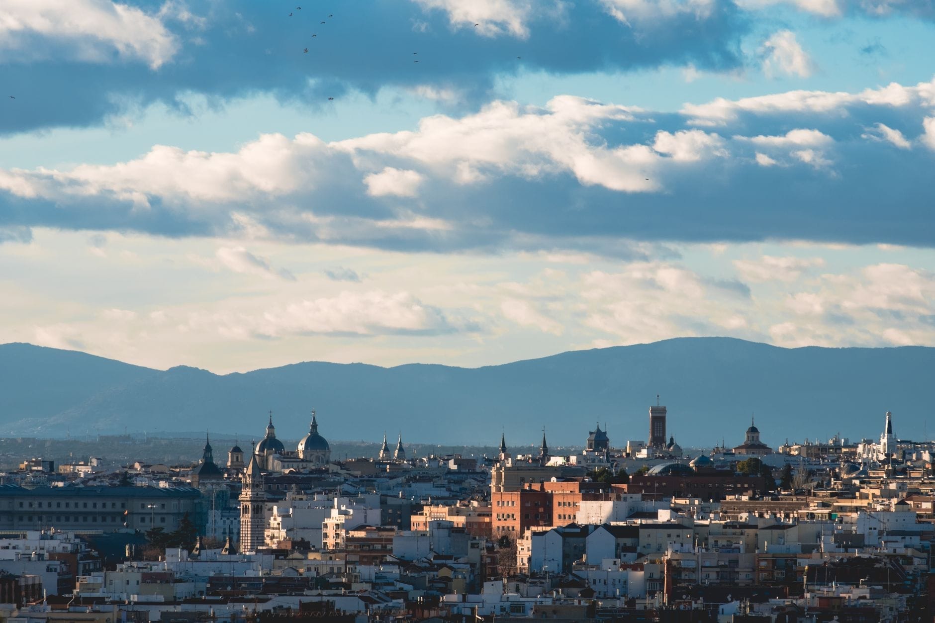 Vista panorâmica de Madrid ao entardecer, mostrando uma mistura de cúpulas históricas, torres e edifícios modernos com montanhas ao fundo sob um céu dramático.