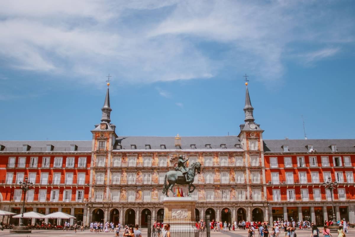 Weitläufige Ansicht der Plaza Mayor in Madrid mit ihren eleganten roten Gebäuden aus der Habsburgerzeit, zwei Türmen, einer zentralen Reiterstatue und Besuchern, die den historischen Platz genießen.