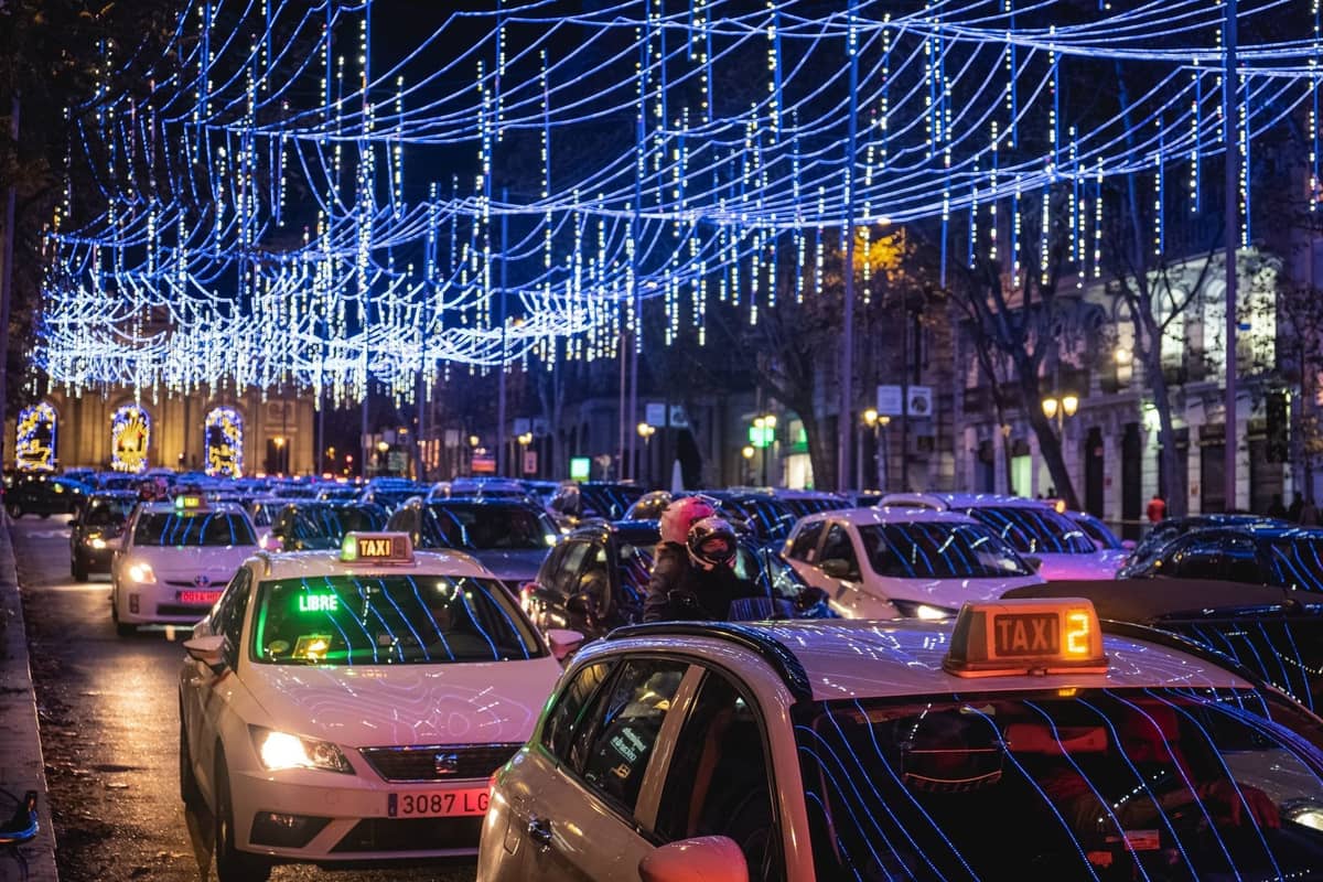 Nächtliche Straßenszene in Madrid mit blauen Weihnachtslichtern, die oben gespannt sind, und Autos, darunter Taxis, im Verkehr unter den festlichen Illuminationen.