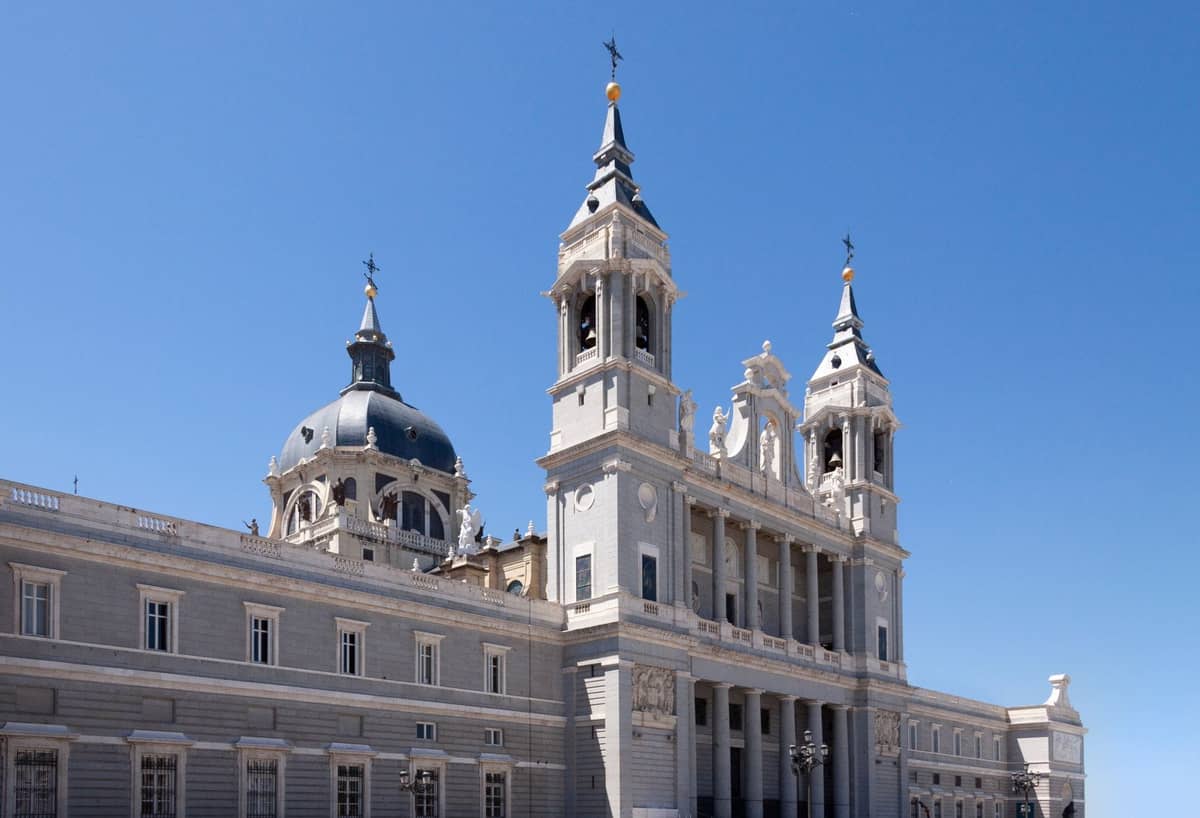 Blick auf die Catedral de la Almudena in Madrid mit ihrer Kuppel, den Zwillingstürmen und der verzierten Fassade vor einem klaren blauen Himmel.