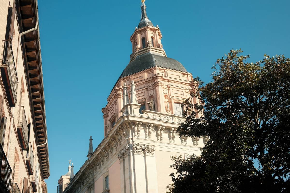 Sonnenbeschienenes Äußere einer historischen Kirche in Madrid mit einer Barockuppel, von der Straße aus unter blauem Himmel betrachtet.