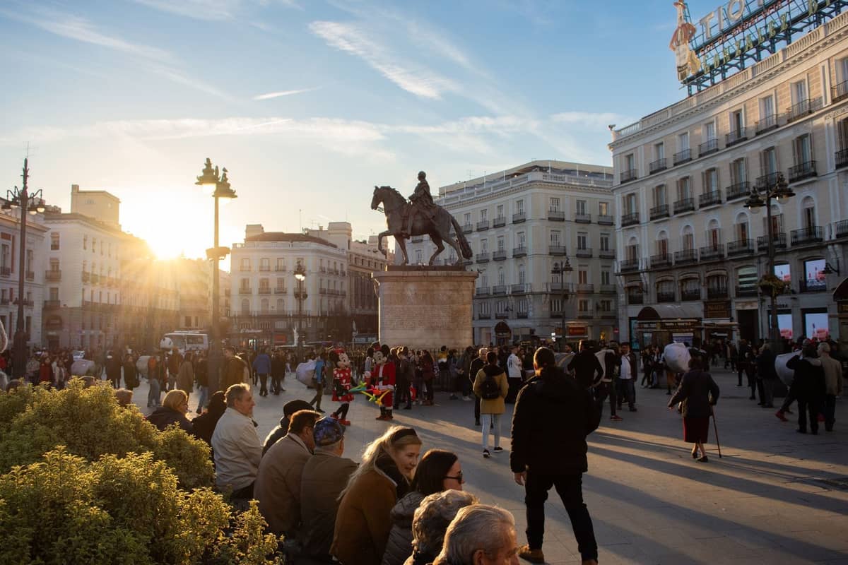 Una bulliciosa Puerta del Sol en Madrid con personas caminando, una estatua de un hombre a caballo y edificios históricos bajo el sol de la tarde.