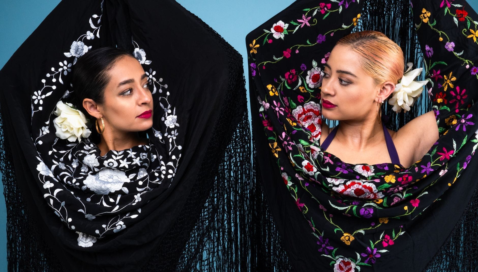 Duas dançarinas de flamenco em xales bordados tradicionais se encaram diante de um fundo azul, evocando fortemente a cultura espanhola e a arte apaixonada do flamenco.