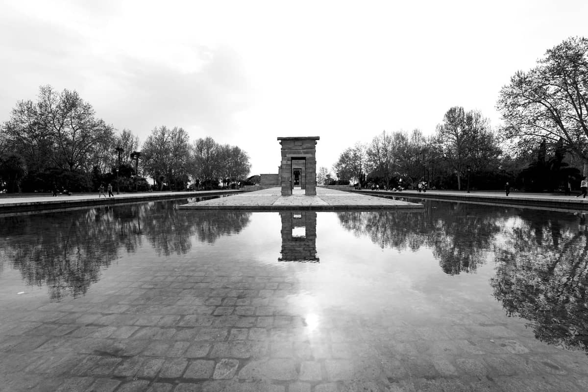 Schwarz-Weiß-Foto des Tempels von Debod, der sich in einem Teich spiegelt, mit Bäumen und Abendhimmel im Hintergrund.