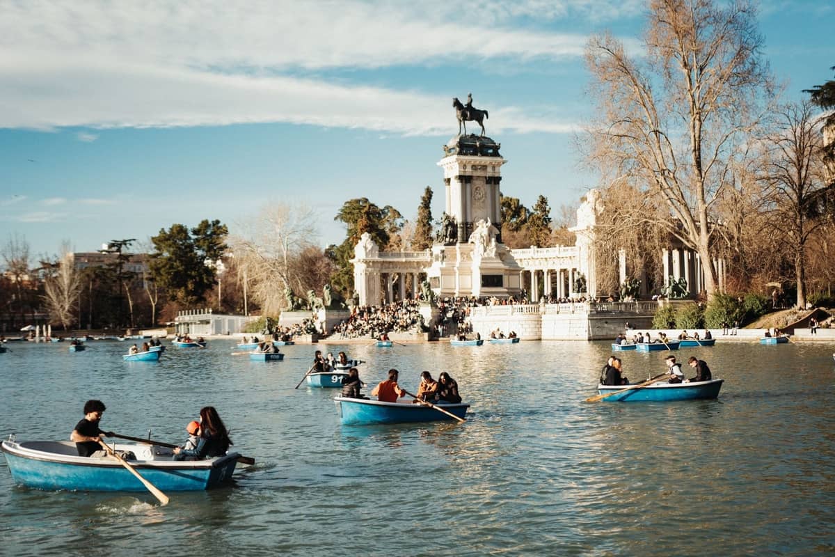 Paare und Gruppen rudern in blauen Booten auf dem großen See im Madrider Parque del Retiro, im Hintergrund das imposante Denkmal Alfonso XII.