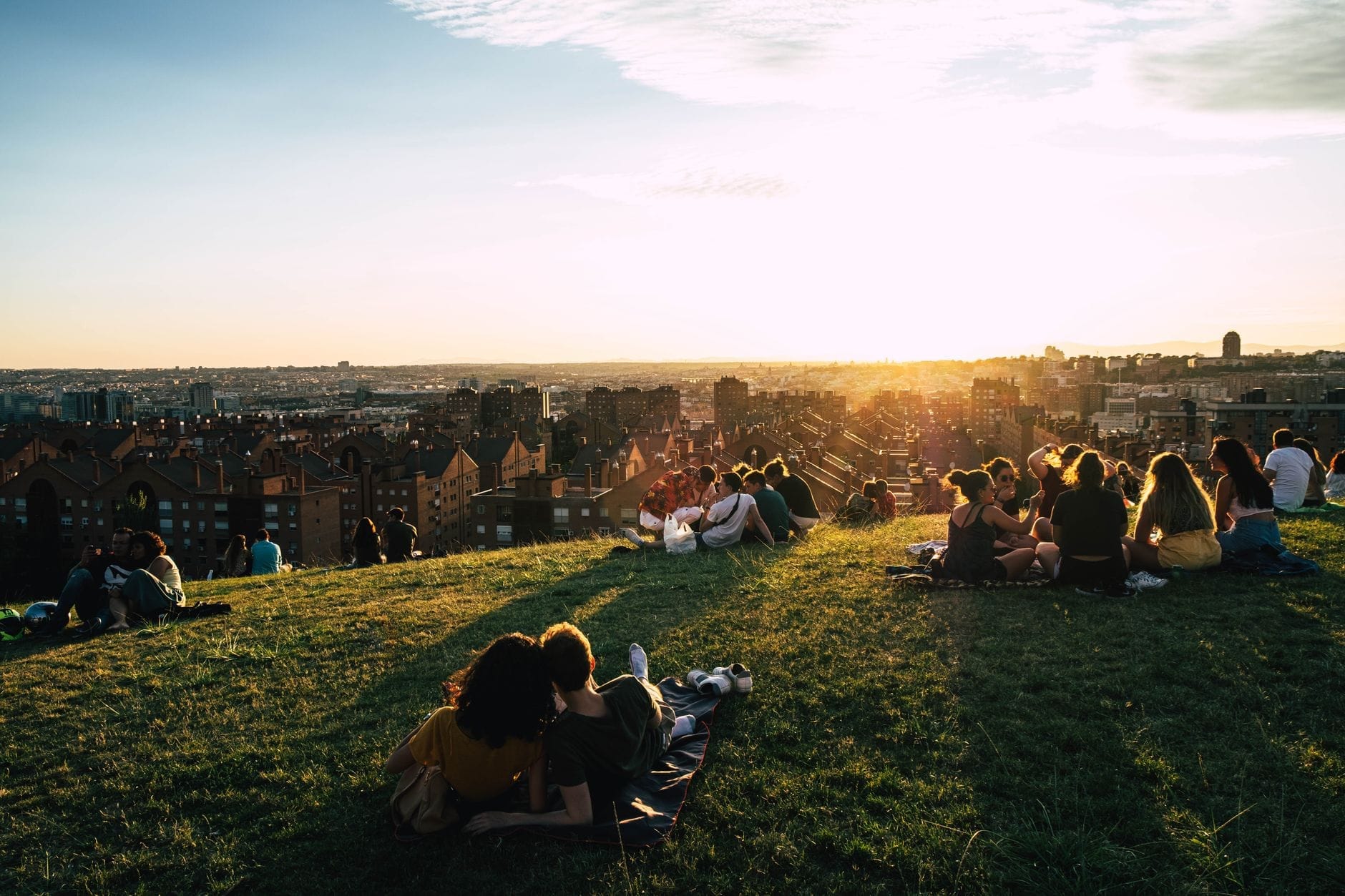 Casal relaxando em uma colina gramada ao pôr do sol com o skyline de Madri ao fundo, banhado por uma luz dourada e quente em uma atmosfera tranquila e romântica.