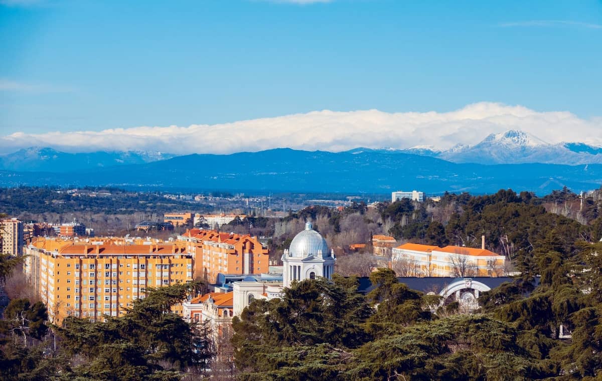 Veduta nitida degli edifici di Madrid con un monumento a cupola bianca e le montagne innevate della Sierra de Guadarrama sullo sfondo sotto un cielo azzurro.