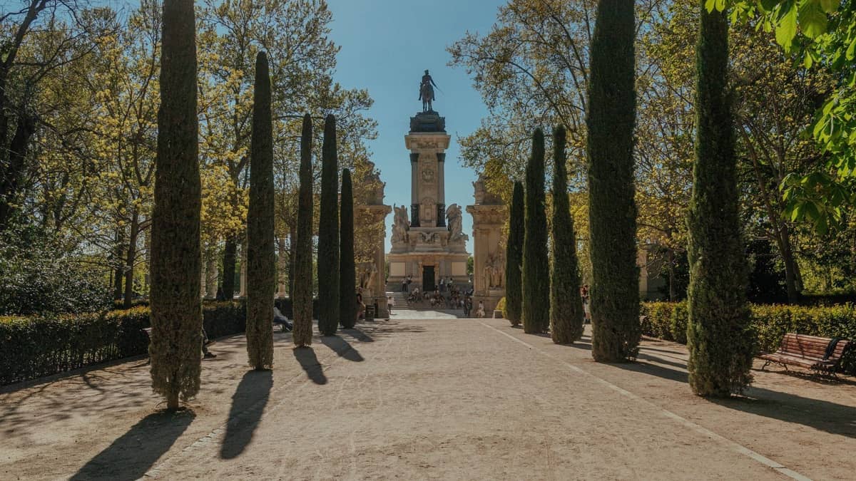 Un vialetto soleggiato in un parco fiancheggiato da alti cipressi che conduce a un grande monumento in uno spazio verde centrale a Madrid.