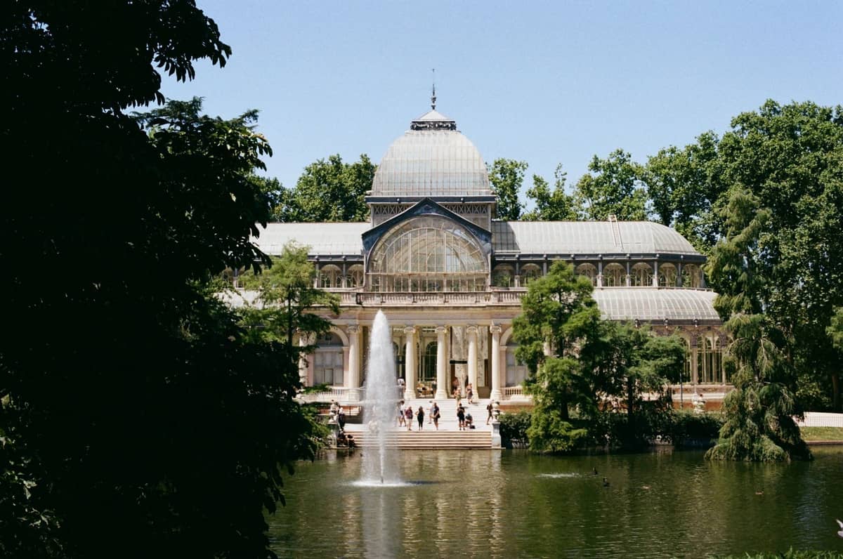 Der Palacio de Cristal im Parque del Retiro in Madrid mit einem See, einem Brunnen und üppigen grünen Bäumen im Frühlingslicht.