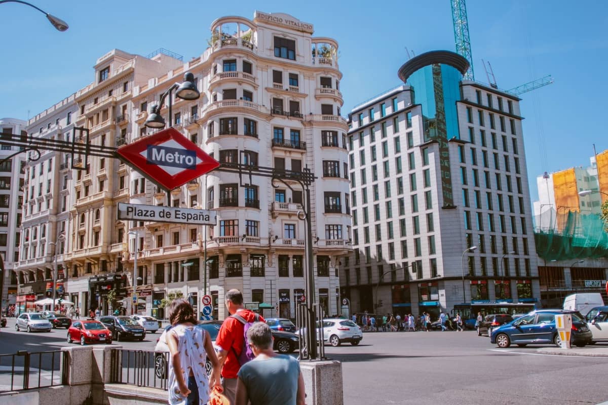 Menschen steigen in die U-Bahn-Station Plaza de España im Zentrum von Madrid hinab, mit Stadtgebäuden und Verkehr an einem sonnigen Tag.