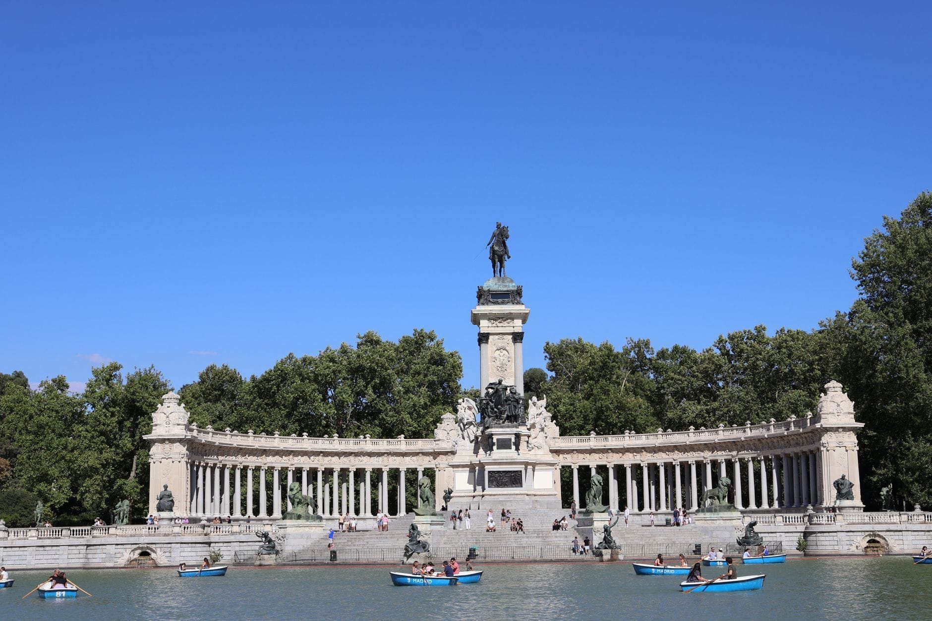 Dia de verão ensolarado no Parque El Retiro, em Madrid, com pessoas passeando de barco no lago em frente ao grande monumento de pedra rodeado de árvores e céu azul.