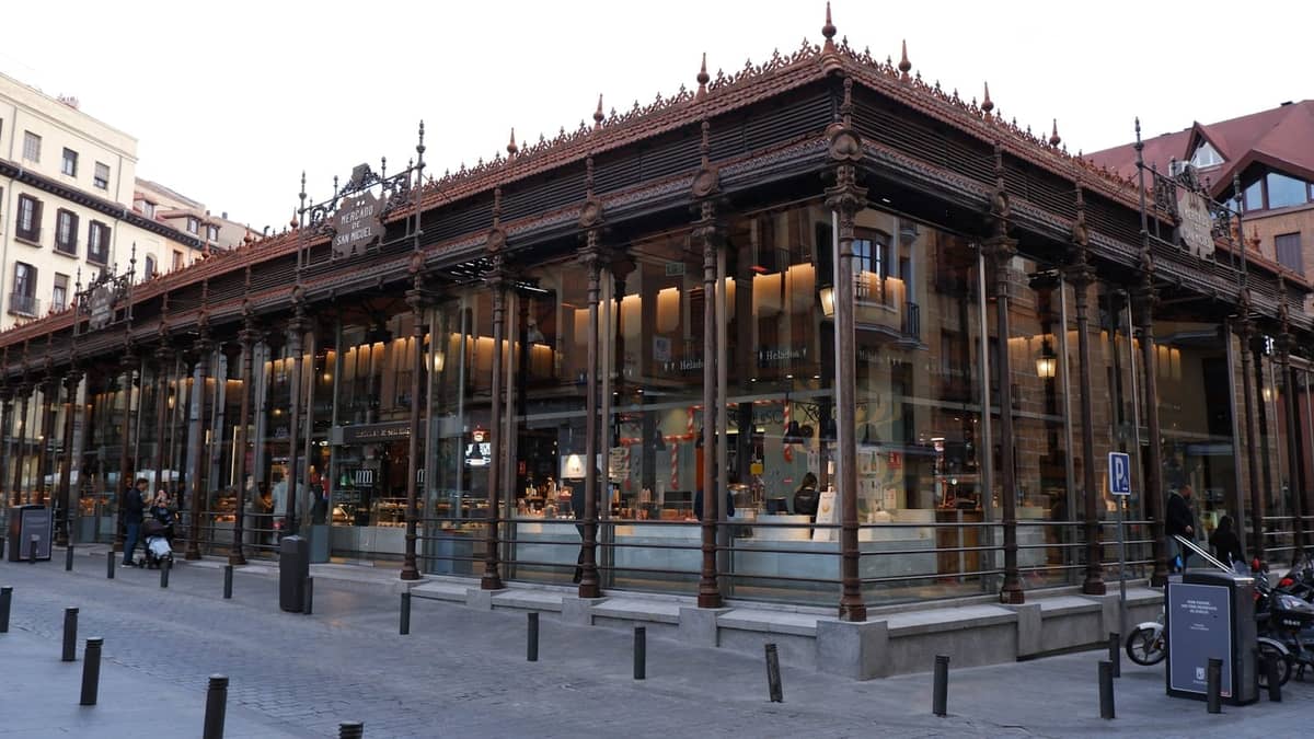 Außenansicht des Mercado de San Miguel in Madrid, eine lebhafte Markthalle aus Glas und Eisen, bekannt für ihre Essensstände und Tapas, mit sichtbaren Menschen im Inneren.